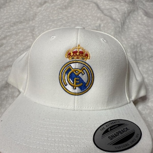 Può includere: Cappellino da baseball bianco con un logo del Real Madrid ricamato sul davanti. Il logo presenta una corona dorata, uno stemma bianco e blu e la lettera "F" in oro. Il cappellino ha un adesivo nero e argento sul retro che dice "The Authentic Yupoong Snapback Since 1974."