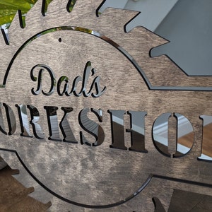 K&ouml;nnte beinhalten: Holzschild mit dem ausgeschnittenen Schriftzug "Dad's WORKSHOP", umrahmt von einem S&auml;geblatt-Design. Das Schild besteht aus dunkel gebeiztem Holz, wobei der Text in einer helleren Farbe gehalten ist. Ein dekoratives St&uuml;ck f&uuml;r die Werkstatt.