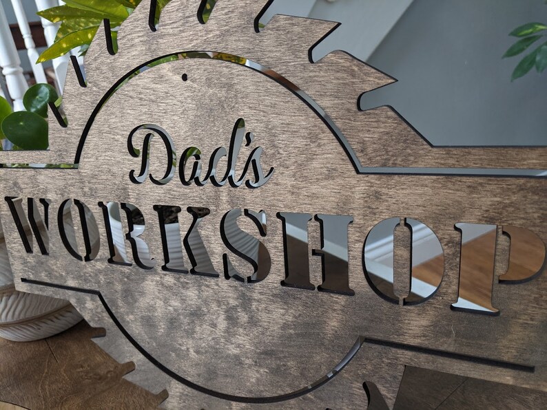 K&ouml;nnte beinhalten: Holzschild mit dem ausgeschnittenen Schriftzug "Dad's WORKSHOP", umrahmt von einem S&auml;geblatt-Design. Das Schild besteht aus dunkel gebeiztem Holz, wobei der Text in einer helleren Farbe gehalten ist. Ein dekoratives St&uuml;ck f&uuml;r die Werkstatt.