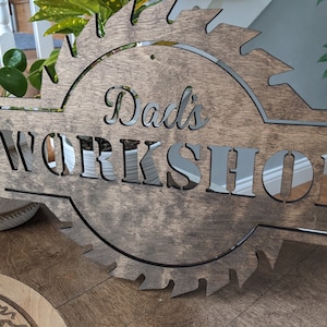 K&ouml;nnte beinhalten: Holzschild mit dem Text "Dad's WORKSHOP" in einem dekorativen S&auml;geblatt-Design ausgeschnitten. Das Schild besteht aus dunklem Braunholz und hat eine reflektierende silberne R&uuml;ckseite f&uuml;r die Buchstaben.