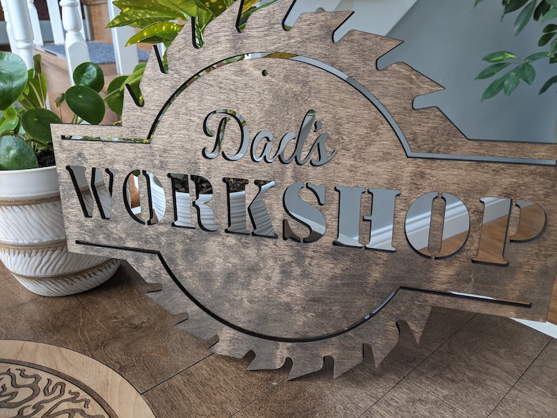 K&ouml;nnte beinhalten: Holzschild mit dem Text "Dad's WORKSHOP" in einem dekorativen S&auml;geblatt-Design ausgeschnitten. Das Schild besteht aus dunklem Braunholz und hat eine reflektierende silberne R&uuml;ckseite f&uuml;r die Buchstaben.