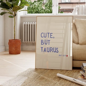 Può includere: Un poster incorniciato bianco con il testo "Cute, But Taurus" in lettere blu e un piccolo cuore rosso con il testo "But Cute" sotto. Il poster è appoggiato a un divano beige in una stanza con una grande finestra e una pianta in vaso.