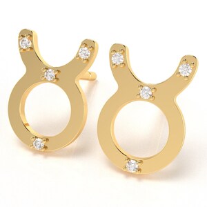 May include: A pair of gold-tone earrings shaped like the Taurus zodiac sign, each with five small clear cubic zirconia stones.