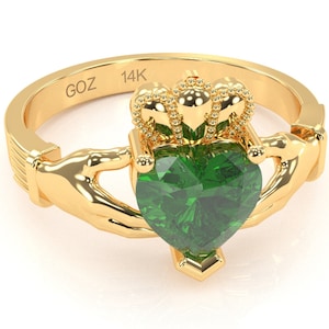 Puede incluir: Un anillo Claddagh de oro de 14 quilates con una piedra preciosa verde en forma de corazón. El anillo presenta dos manos que sujetan un corazón con una corona en la parte superior.