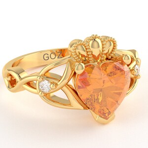 Könnte beinhalten: Ein goldener Ring mit einem herzförmigen orangefarbenen Edelstein und zwei kleineren Diamanten. Der Ring hat ein keltisches Knotendesign und eine Krone oben.