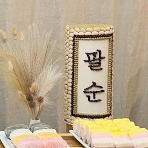 Op de afbeelding: Een presentatie van traditionele Koreaanse desserts, waaronder kleurrijke rijstcakes en een decoratieve pilaar met Koreaanse tekst. De pilaar is versierd met kleine, lichtgekleurde objecten en donkerbruine accenten. De desserts zijn gerangschikt op witte borden.