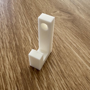 May include: A white, L-shaped 3D-printed object with a circular hole in the vertical section. The object is made of a solid, opaque material and sits on a wood-grain surface.