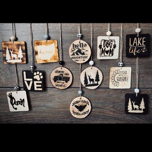 May include: A collection of 12 wooden ornaments with various designs and sayings. The ornaments are all round or square and have a rustic, wood-burned look. Some of the sayings include "home", "hello adventure", "dog mom", "lake life", "love", "country roads take me home", "you are my sunshine", and "queen".