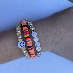 Op de afbeelding: Drie kralenarmbanden gestapeld op een pols. De bovenste armband heeft witte kralen met het woord "FALL" gespeld in gouden letters. De middelste armband heeft oranje, rode en zwarte kralen. De onderste armband heeft witte kralen met een smiley-bedel.