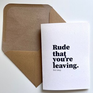 May include: A white greeting card with navy blue text reading "Rude that you're leaving." and "But okay." in a smaller font, alongside a brown envelope. A simple, modern design.