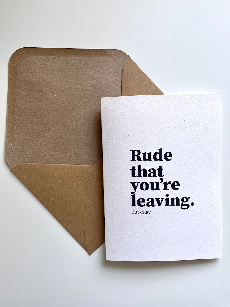 May include: A white greeting card with navy blue text reading "Rude that you're leaving." and "But okay." in a smaller font, alongside a brown envelope. A simple, modern design.