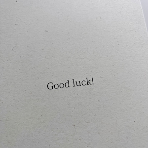 May include: An off-white textured card with the words "Good luck!" printed in black. The card's design is simple and elegant, suitable for various occasions.