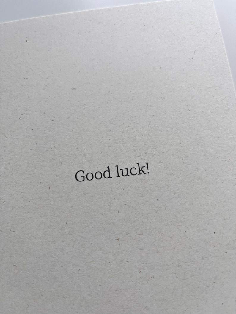 May include: An off-white textured card with the words "Good luck!" printed in black. The card's design is simple and elegant, suitable for various occasions.