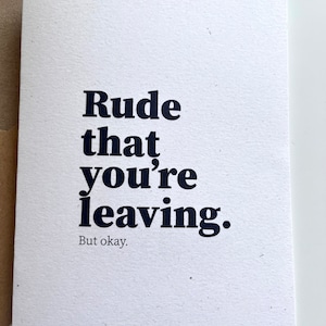May include: A white card with the words "Rude that you're leaving. But okay." printed in a dark blue, bold font. The card is rectangular and made of textured paper. A brown envelope is partially visible in the background.