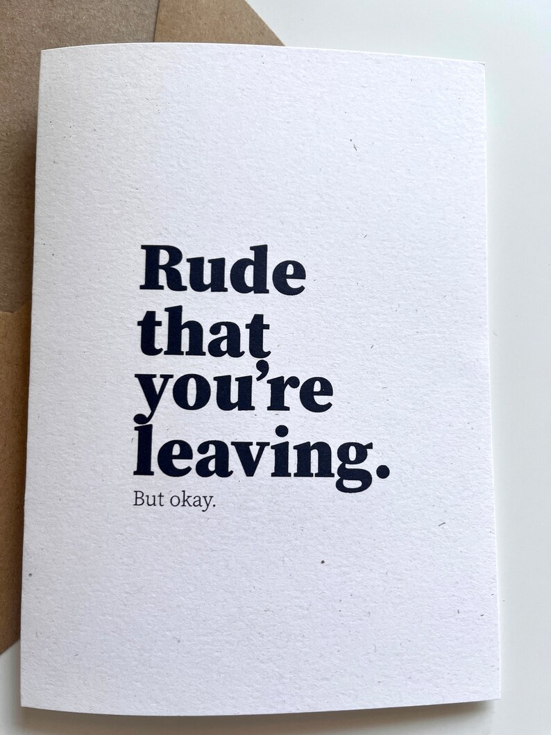May include: A white card with the words "Rude that you're leaving. But okay." printed in a dark blue, bold font. The card is rectangular and made of textured paper. A brown envelope is partially visible in the background.