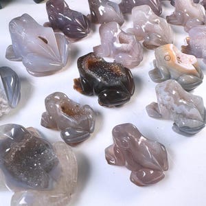 Puede incluir: Una colección de esculturas de ranas de ágata en varios tonos de gris, marrón y blanco. Las ranas están talladas con detalles intrincados y tienen un acabado natural y pulido.