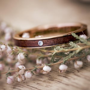 Puede incluir: Un anillo de banda de oro rosa con un solo diamante engastado en una incrustaci&oacute;n de madera. El anillo descansa sobre un lecho de delicadas flores blancas.