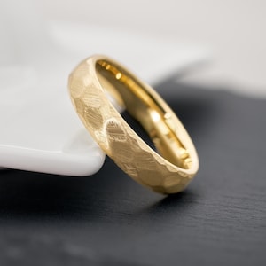 Könnte beinhalten: Ein goldfarbener Ring mit einer strukturierten, gehämmerten Oberfläche. Der Ring ist vor einem kontrastierenden schwarz-weißen Hintergrund platziert, der seinen metallischen Glanz hervorhebt. Das Band hat eine abgerundete Form, und die Innenseite des Rings ist glatt und poliert.