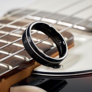 Może przedstawiać: Czarny pierścionek z białym wewnętrznym paskiem i srebrnym wzorem struny, spoczywający na gitarze. Pierścionek ma błyszczące wykończenie i małe logo. Widoczna jest drewniana podstrunnica i struny gitary.