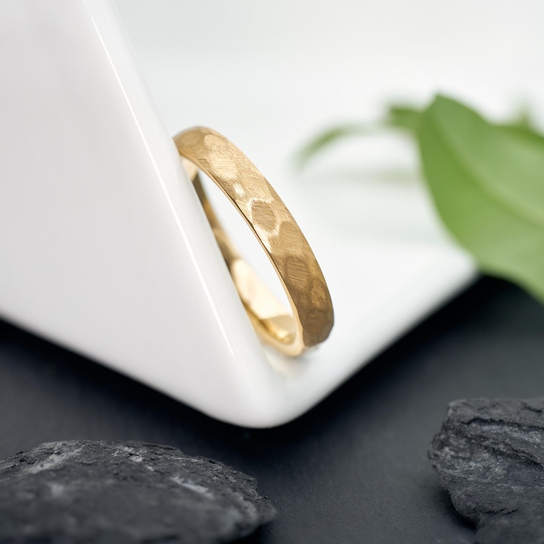 May include: A gold ring with a hammered texture, resting on a white surface. The ring's surface reflects light, showcasing its detailed design. The ring is a piece of jewelry.