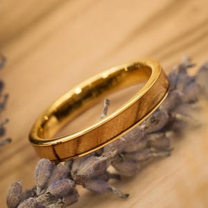Peut inclure: Une bague dorée avec un incrustation en bois. La bague est posée sur un fond flou de lavande et une surface brun clair. Le design de la bague présente une fine bande de bois encastrée dans un métal doré.