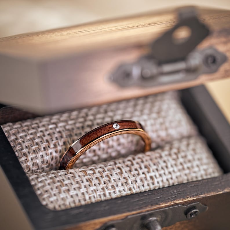 Puede incluir: Una caja de madera para anillos con una incrustaci&oacute;n de madera marr&oacute;n y un anillo de diamante en su interior. El anillo est&aacute; hecho de metal con una incrustaci&oacute;n de madera marr&oacute;n.