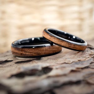 May include: Two wedding rings with a black band and a wood inlay. The rings are made of a dark wood and are set against a textured, brown background. The rings are close together, suggesting a couple.
