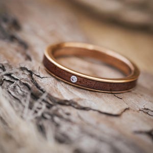 Puede incluir: Una banda de oro rosa con una fina incrustaci&oacute;n de madera oscura y un peque&ntilde;o diamante engastado en el metal.