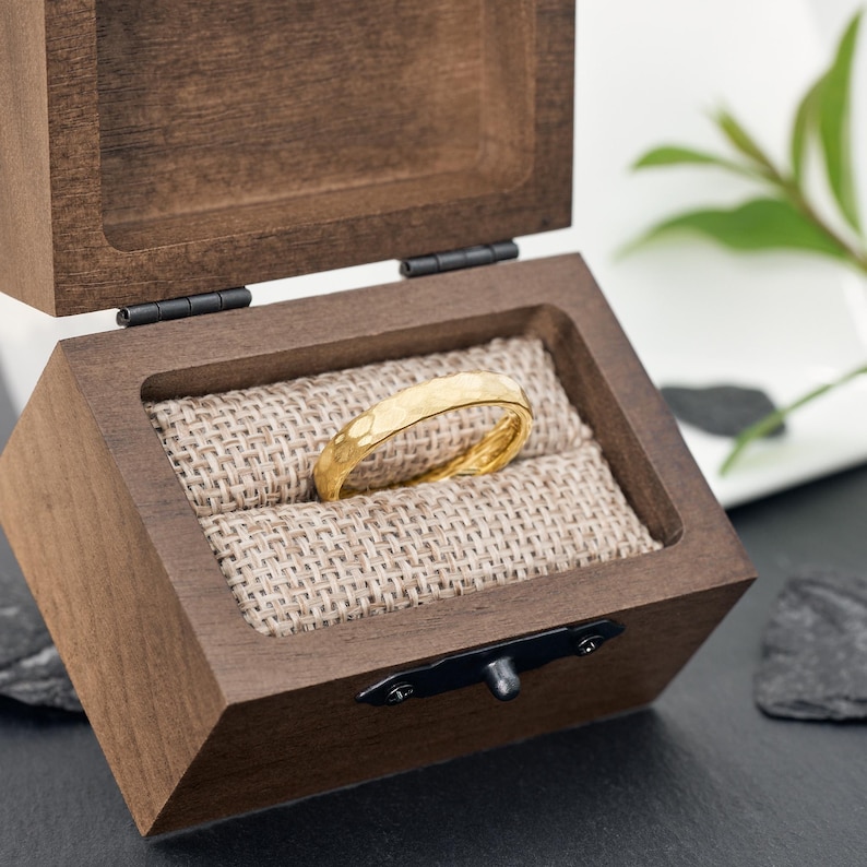 May include: A gold ring with a hammered texture rests inside a wooden box lined with burlap. The box is open, revealing the ring's intricate design. The ring is a warm gold color, and the box is a dark brown.