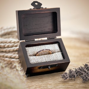 Puede incluir: Una caja de madera para anillos con una tapa con bisagras, abierta para revelar una fina banda dorada con un solo diamante engastado en el centro. El anillo descansa sobre un coj&iacute;n de tela beige dentro de la caja.