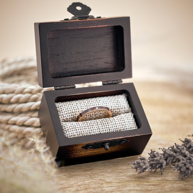 Puede incluir: Una caja de madera para anillos con una tapa con bisagras, abierta para revelar una fina banda dorada con un solo diamante engastado en el centro. El anillo descansa sobre un coj&iacute;n de tela beige dentro de la caja.