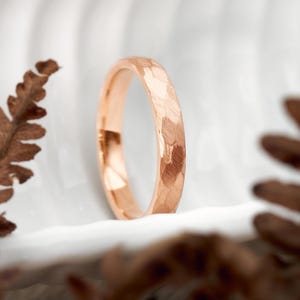 Könnte beinhalten: Ein roségoldener Ring mit gehammerter Textur. Der Ring ist vor einem weißen Hintergrund mit verschwommenem braunen Laub auf beiden Seiten platziert. Die Oberfläche des Rings reflektiert Licht und hebt das strukturierte Design hervor.