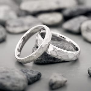 May include: Two silver rings with a textured, hammered finish. The rings are displayed on a gray surface with several gray stones in the background. The rings have a simple, elegant design.