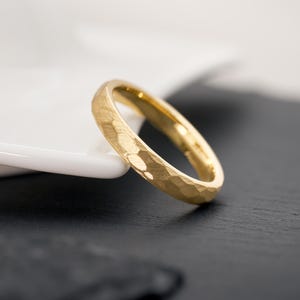 May include: A gold ring with a textured, hammered finish. The ring is a simple band design, with a slightly rounded profile. The ring is set against a black and white background, highlighting its golden color.