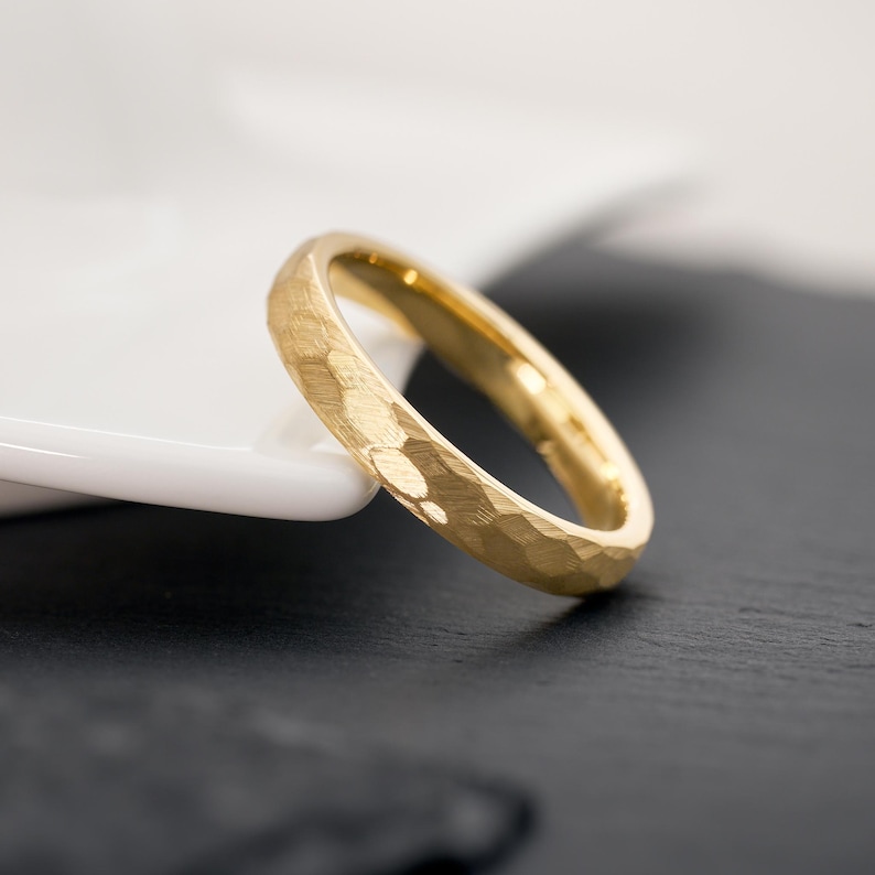 May include: A gold ring with a textured, hammered finish. The ring is a simple band design, with a slightly rounded profile. The ring is set against a black and white background, highlighting its golden color.