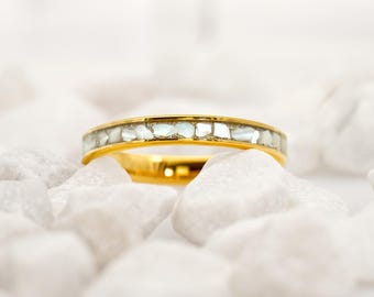 May include: A gold ring with a band inlaid with small, irregular, light-colored stones. The ring is set against a backdrop of white, rounded stones, creating a contrast of textures and colors. The ring's polished gold reflects the light.