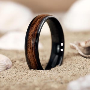 Puede incluir: Primer plano de un anillo con un centro de veta de madera oscura y un exterior negro brillante. El anillo se encuentra sobre un fondo arenoso con conchas marinas, destacando su estética natural. El diseño del anillo es simple y elegante.