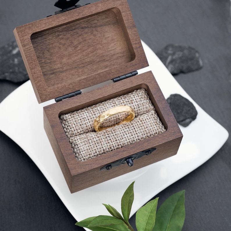 May include: A gold ring with a textured surface rests inside an open, dark wooden ring box. The box is lined with burlap and sits on a white plate. The ring box is secured with a black clasp.