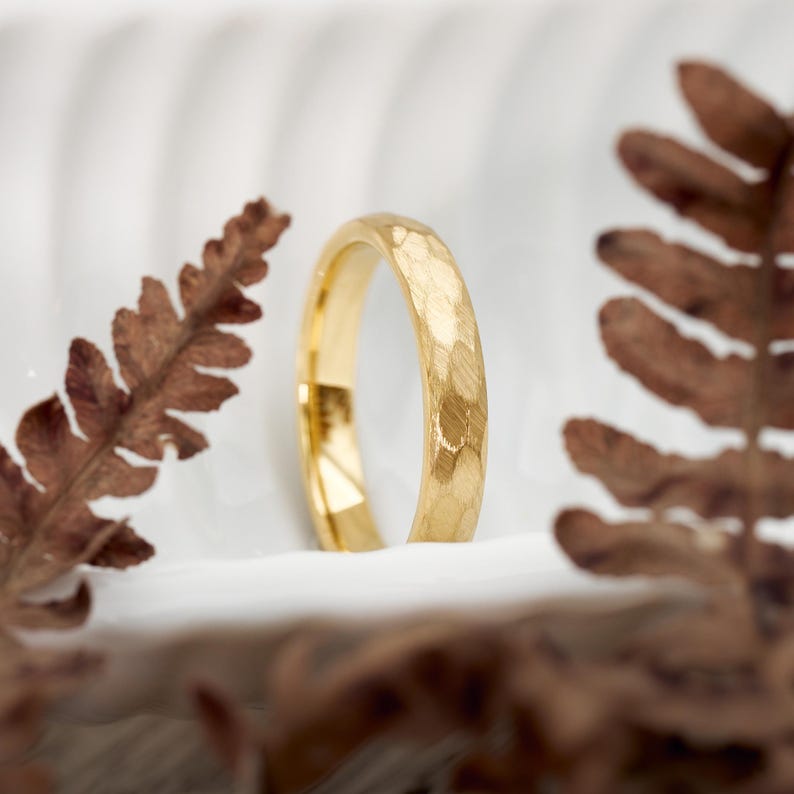 May include: A gold ring with a textured surface stands upright against a white background, flanked by brown fern leaves. The ring's surface has a hammered appearance, catching the light. The ring is a piece of jewelry.