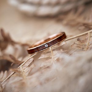 Puede incluir: Un fino anillo de oro con un peque&ntilde;o diamante engastado en una incrustaci&oacute;n de madera. El anillo descansa sobre un lecho de helechos secos.