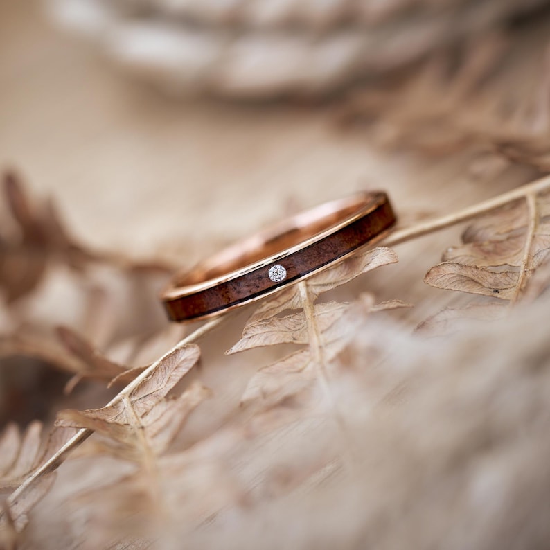 Puede incluir: Un fino anillo de oro con un peque&ntilde;o diamante engastado en una incrustaci&oacute;n de madera. El anillo descansa sobre un lecho de helechos secos.