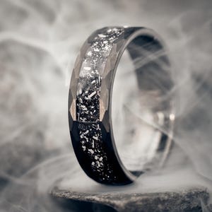Op de afbeelding: Een zwarte ring met een facetontwerp en een zilveren inleg. De ring staat tegen een rokerige achtergrond, die de textuur en details benadrukt. De ring lijkt van metaal te zijn en heeft een gepolijste afwerking.