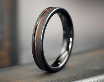May include: A black ring with a brushed metal finish and a thin, orange-brown wood inlay. The ring has a polished, reflective edge. The ring is shown on a textured, rusty metal surface.