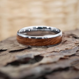 Könnte beinhalten: Nahaufnahme eines Eherings mit silbernem Band und Holzeinlage. Der Ring ist vor einem strukturierten, braunen Hintergrund platziert, der den Kontrast zwischen dem glänzenden Metall und der natürlichen Holzmaserung hervorhebt.