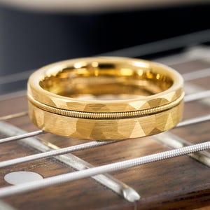May include: A gold-colored ring with a textured, geometric design and a guitar string inlay. The ring is set against a wooden guitar fretboard, showcasing its musical theme. The ring's band has a width of approximately 0.3 inches.