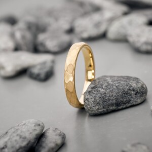 May include: A gold ring with a textured surface, standing upright on a gray stone. The ring has a hammered design and a polished finish. The background features other gray stones, creating a natural, minimalist aesthetic.