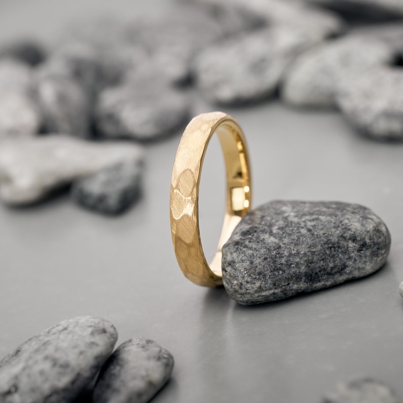 May include: A gold ring with a textured surface, standing upright on a gray stone. The ring has a hammered design and a polished finish. The background features other gray stones, creating a natural, minimalist aesthetic.