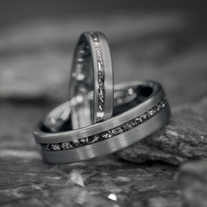 Könnte beinhalten: Zwei silberne Ringe mit strukturierter Oberfläche. Jeder Ring hat ein Band aus dunklem, gesprenkeltem Material. Die Ringe sind gestapelt, wobei einer aufrecht steht und die komplizierten Details des Designs zeigt. Der Hintergrund ist unscharf.