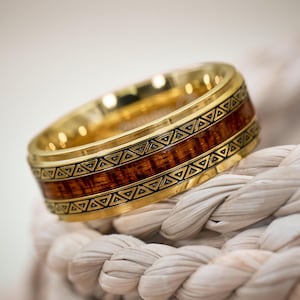 Könnte beinhalten: Ein goldfarbener Ring mit Holzeinlage und geometrischen Details. Der Ring hat ein Band aus dunkelbraunem Holz, das von goldenem Metall mit einem sich wiederholenden Dreiecksmuster umrahmt wird. Der Ring liegt auf einem weißen Seil.