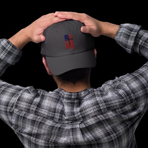 May include: A dark gray baseball cap with an embroidered American flag featuring a red cross in the center.
